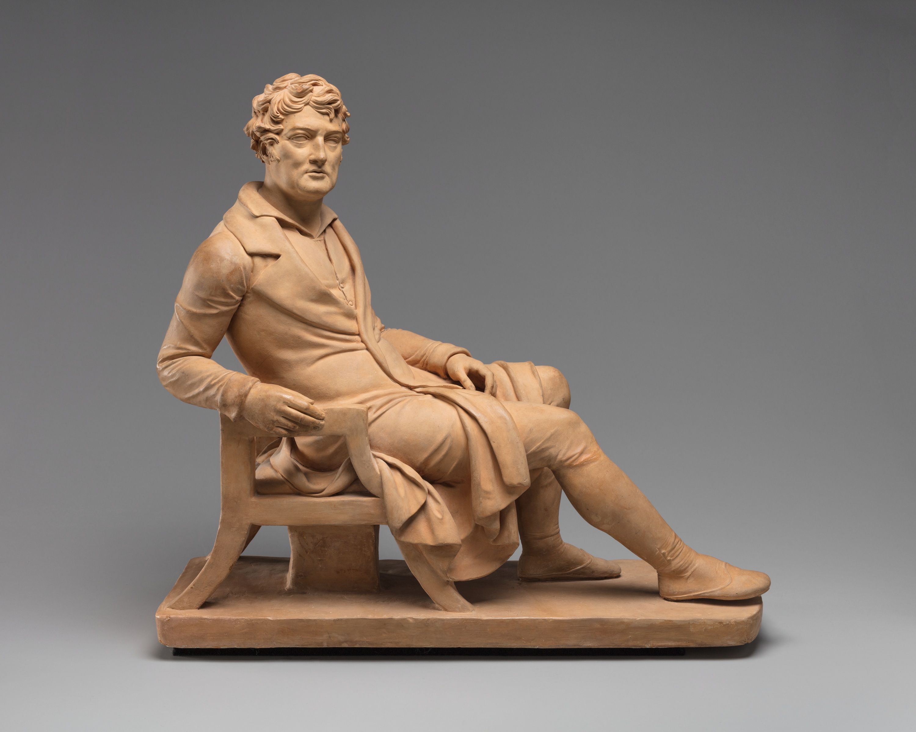 A terracotta statue of a gentleman reclining on a chair.
