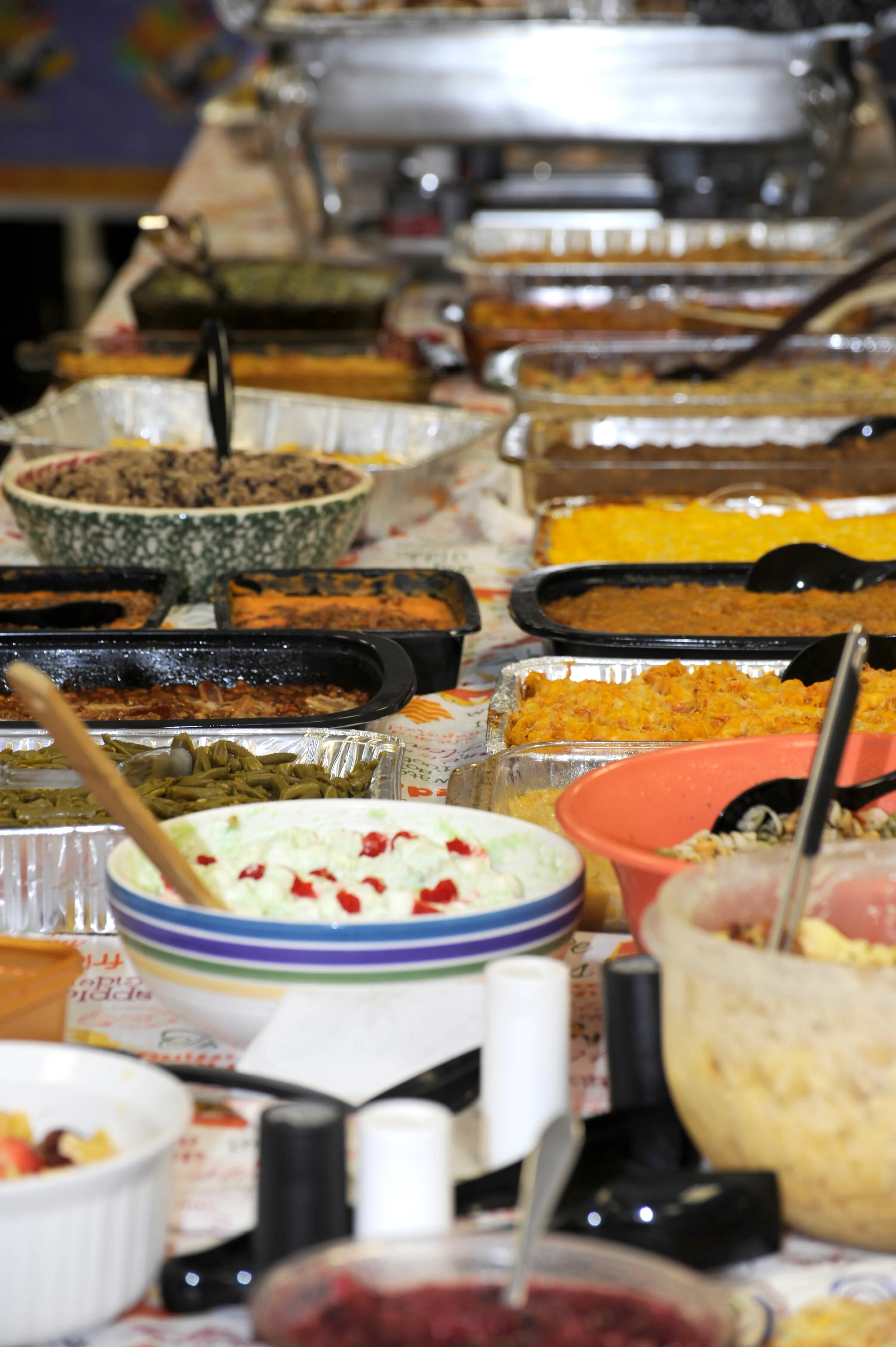 Picture of a Thanksgiving potluck spread as seen from the narrow side of the table. At least 18 different dishes are pictured in various casserole dishes, including what looks like stuffing, macaroni and cheese, sweet potatoes, and green beans.