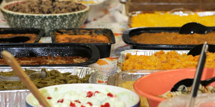 Picture of a Thanksgiving potluck spread as seen from the narrow side of the table. At least 18 different dishes are pictured in various casserole dishes, including what looks like stuffing, macaroni and cheese, sweet potatoes, and green beans.