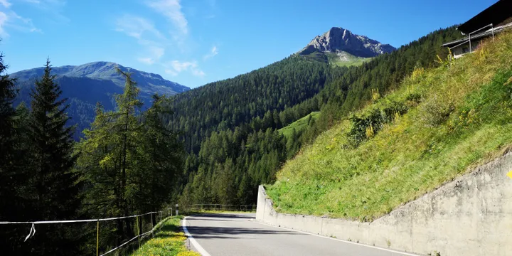 A photo of a curve in a mountain road. There is a ribbon marker along the left side before a steep drop-off. A concrete retaining wall is on the right side.
