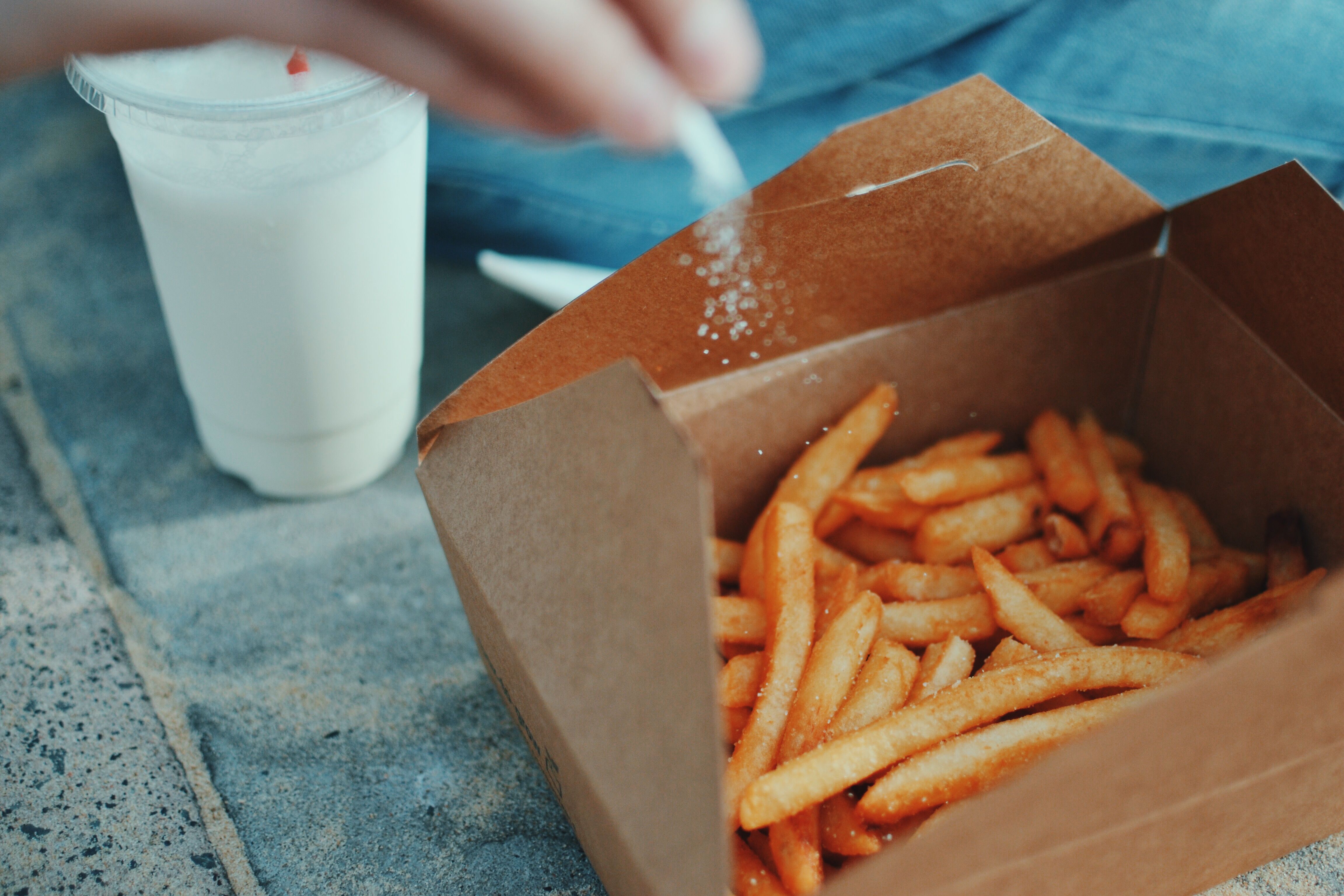 A view of a takeout box filled with french fries. An out-of-focus hand in the foreground is sprinkling salt over them. A white milkshake is in the background.

