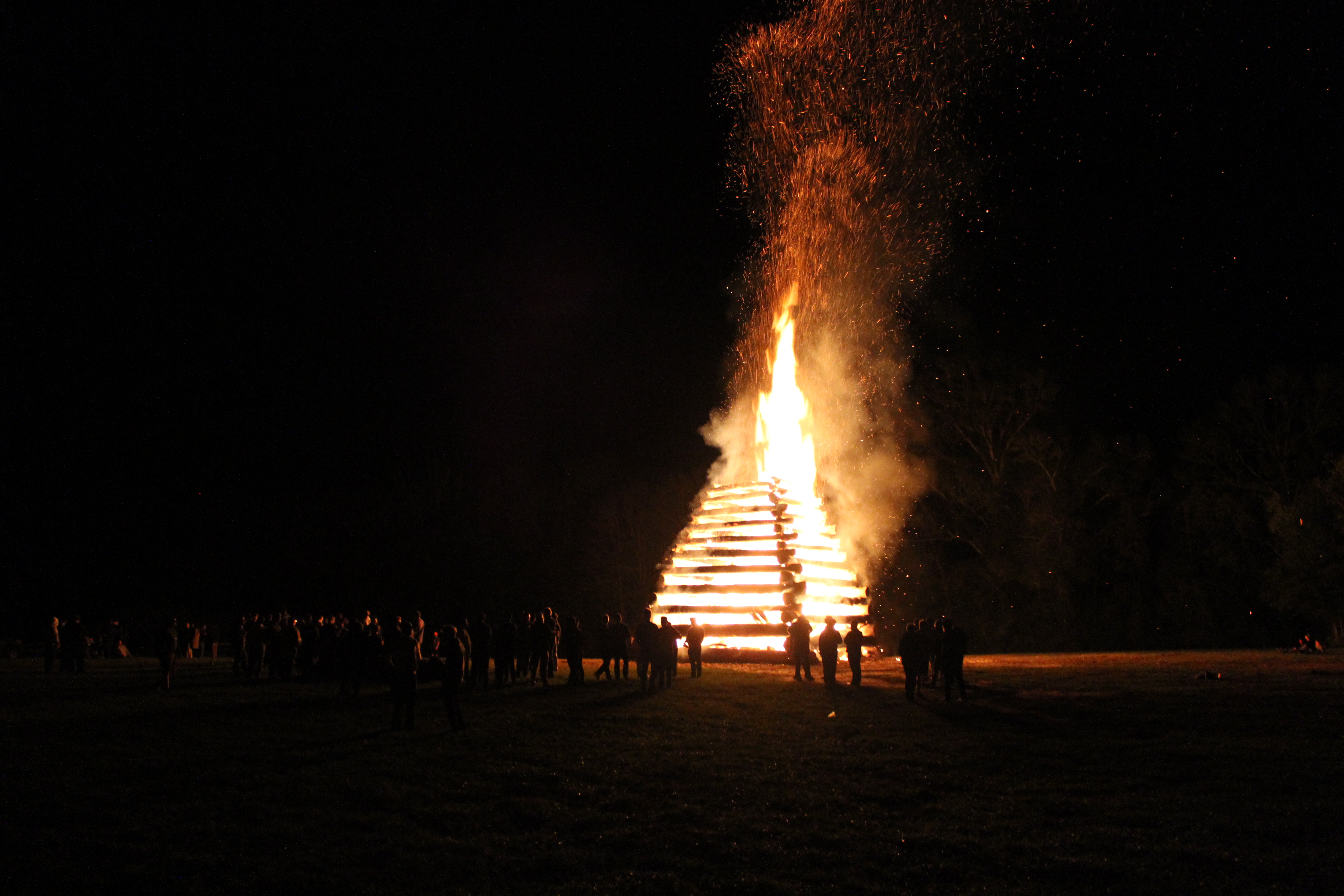 A picture of a bonfire at night. It is a pyramid-like structure standing at least 25 feet high with the flames extending another 15 feet.
